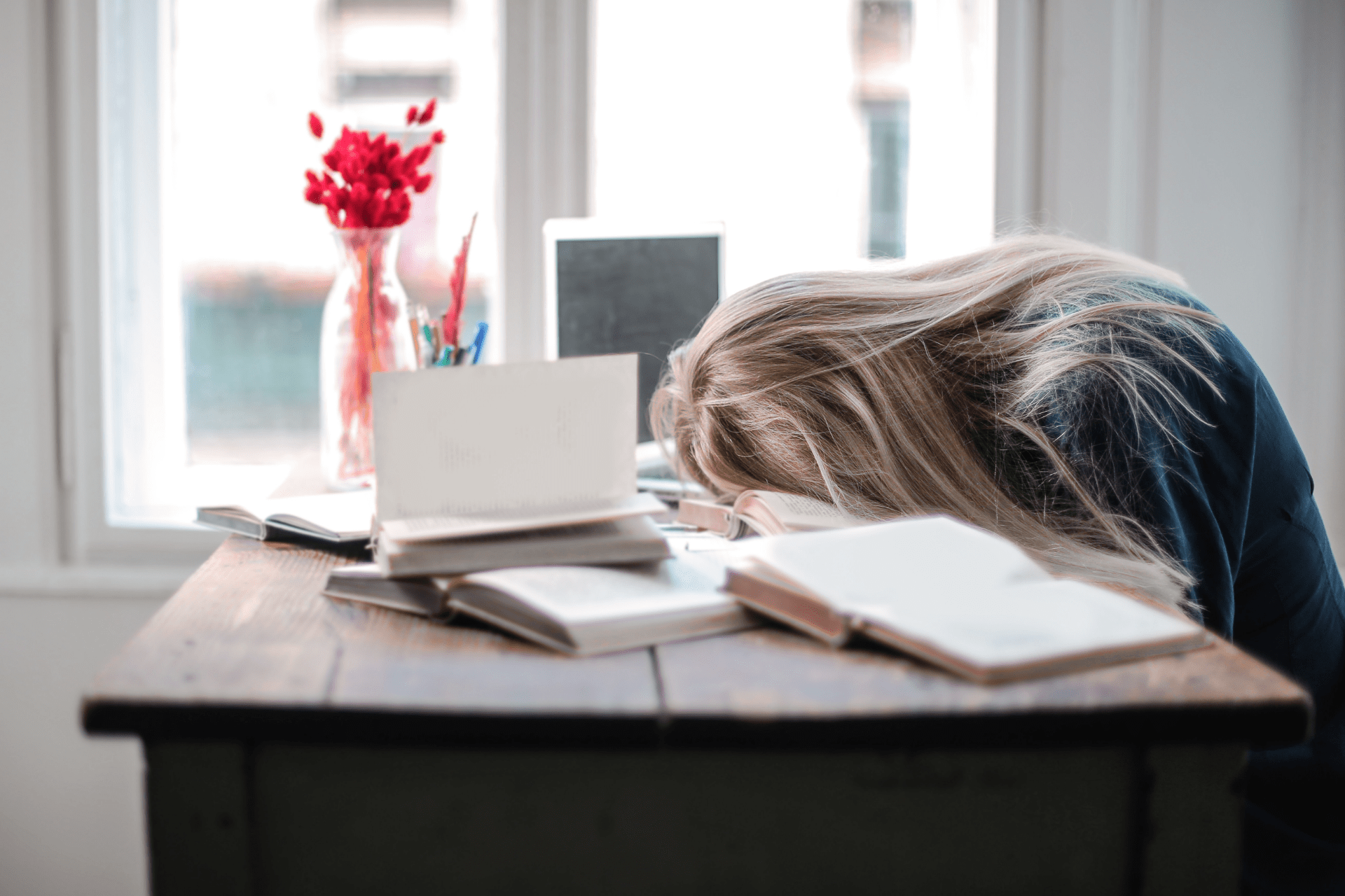 Long-haired blond student asleep with head on desk covered in books and laptop